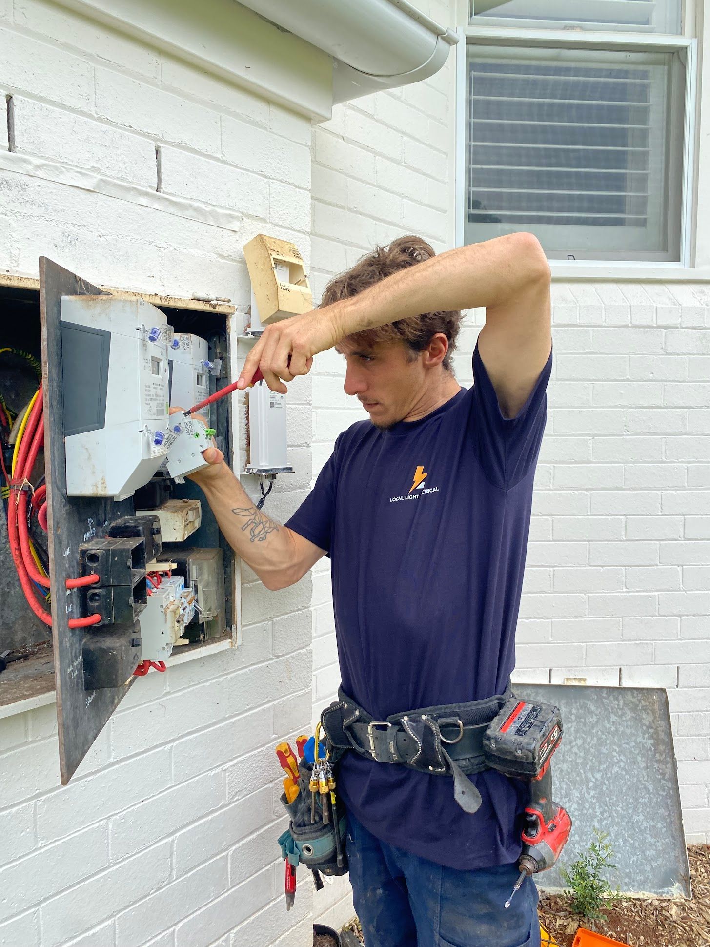 Electrician wiring an open outdoor electrical panel beside a white brick wall.
