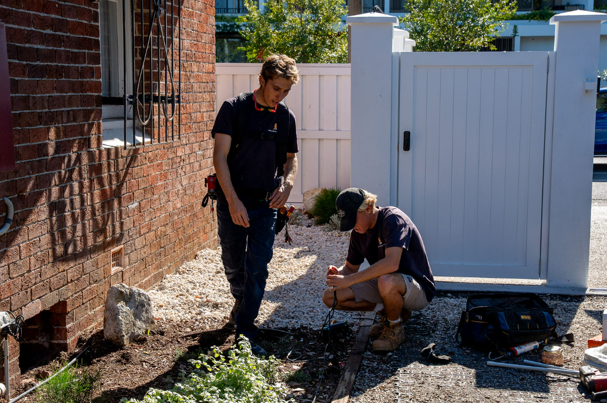 Two People Work Beside a Brick House and White Fence — Local Light Electrical in Suffolk Park, NSW