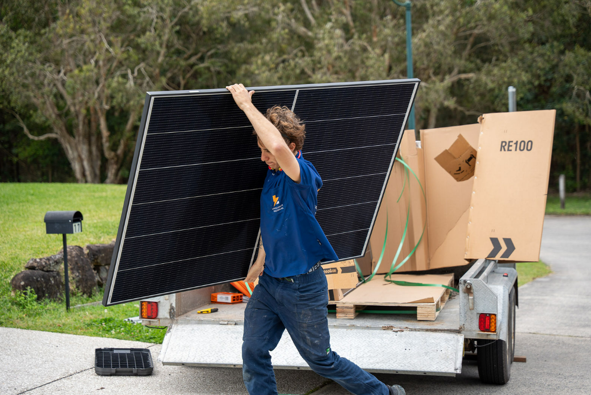 Person Lifting a Large Solar Panel From a Trailer in a Grassy Outdoor Area — Local Light Electrical in Suffolk Park, NSW