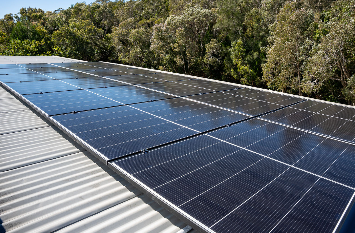 Worker in safety harness standing on solar panels beside a large panel under a bright sky.