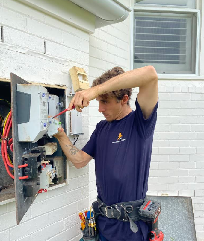 Electrician Wiring an Outdoor Breaker Panel Beside a White Brick Wall — Local Light Electrical in Suffolk Park, NSW