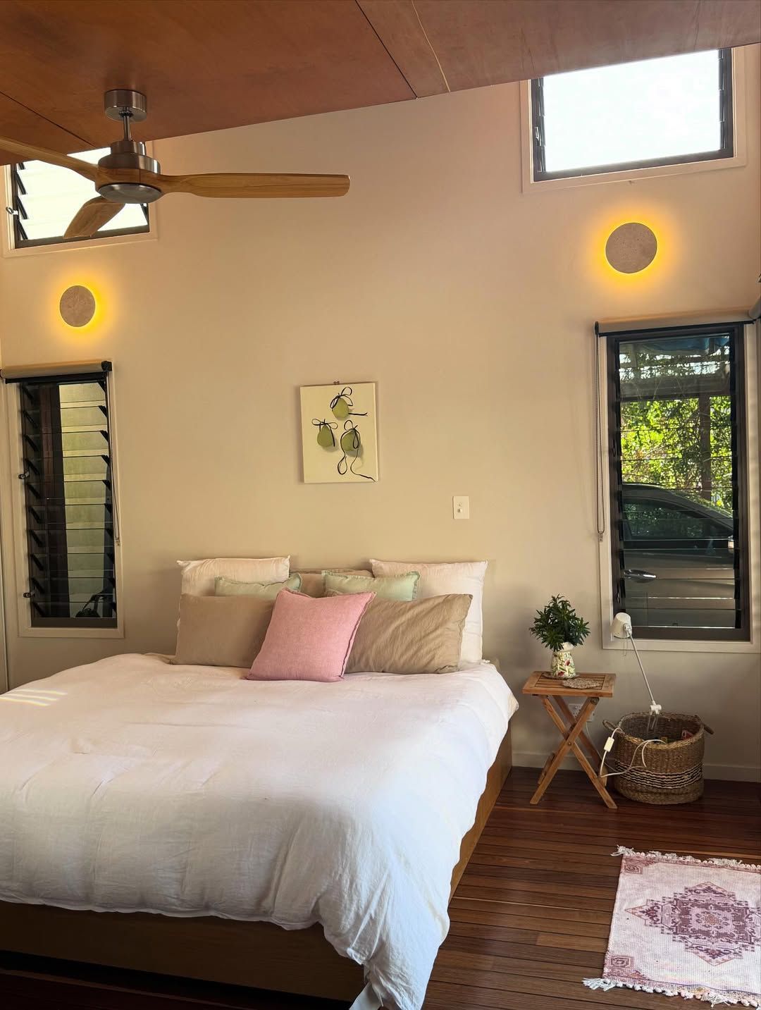 Bright bedroom with white bed, pink pillow, wood ceiling fan, and windows with greenery outside