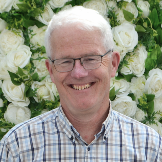 A man wearing glasses and a plaid shirt is smiling in front of a wall of white flowers.