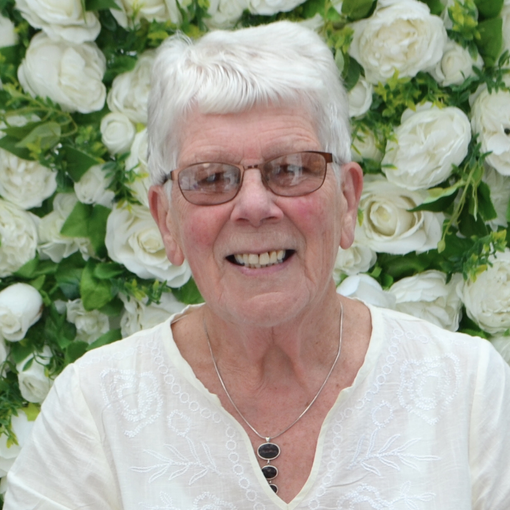 An elderly woman wearing glasses and a necklace smiles in front of a wall of white roses