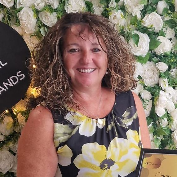 A woman in a floral dress is smiling in front of a wall of flowers.