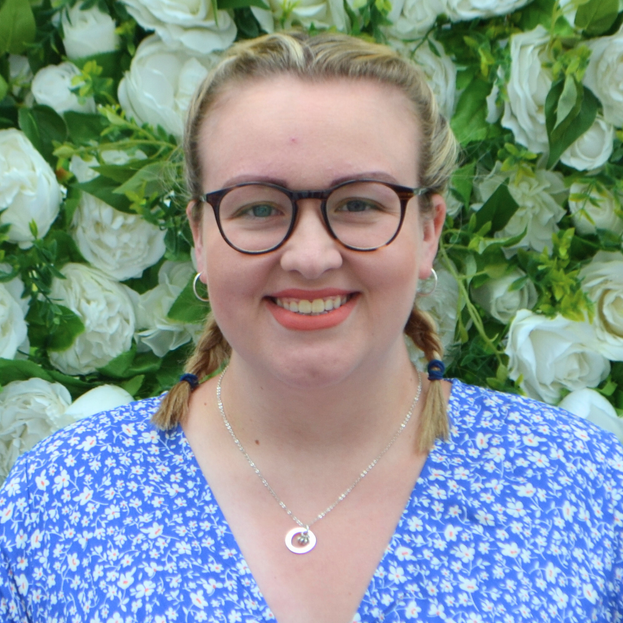 A woman wearing glasses and a necklace is smiling in front of a wall of flowers.