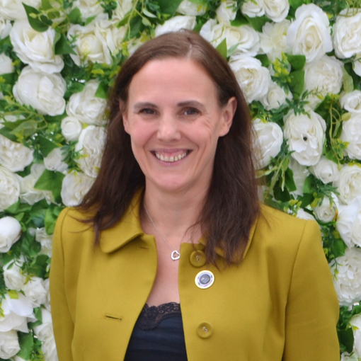 A woman in a yellow jacket is smiling in front of a wall of white flowers