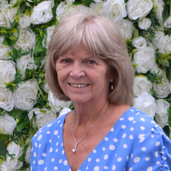 A woman in a blue polka dot dress is smiling in front of a wall of white flowers.