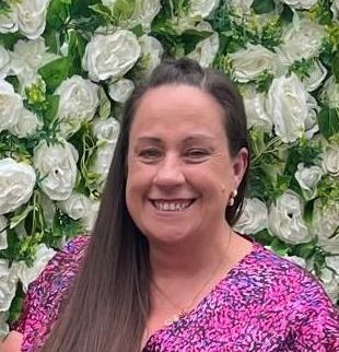 A woman is smiling in front of a wall of white flowers.