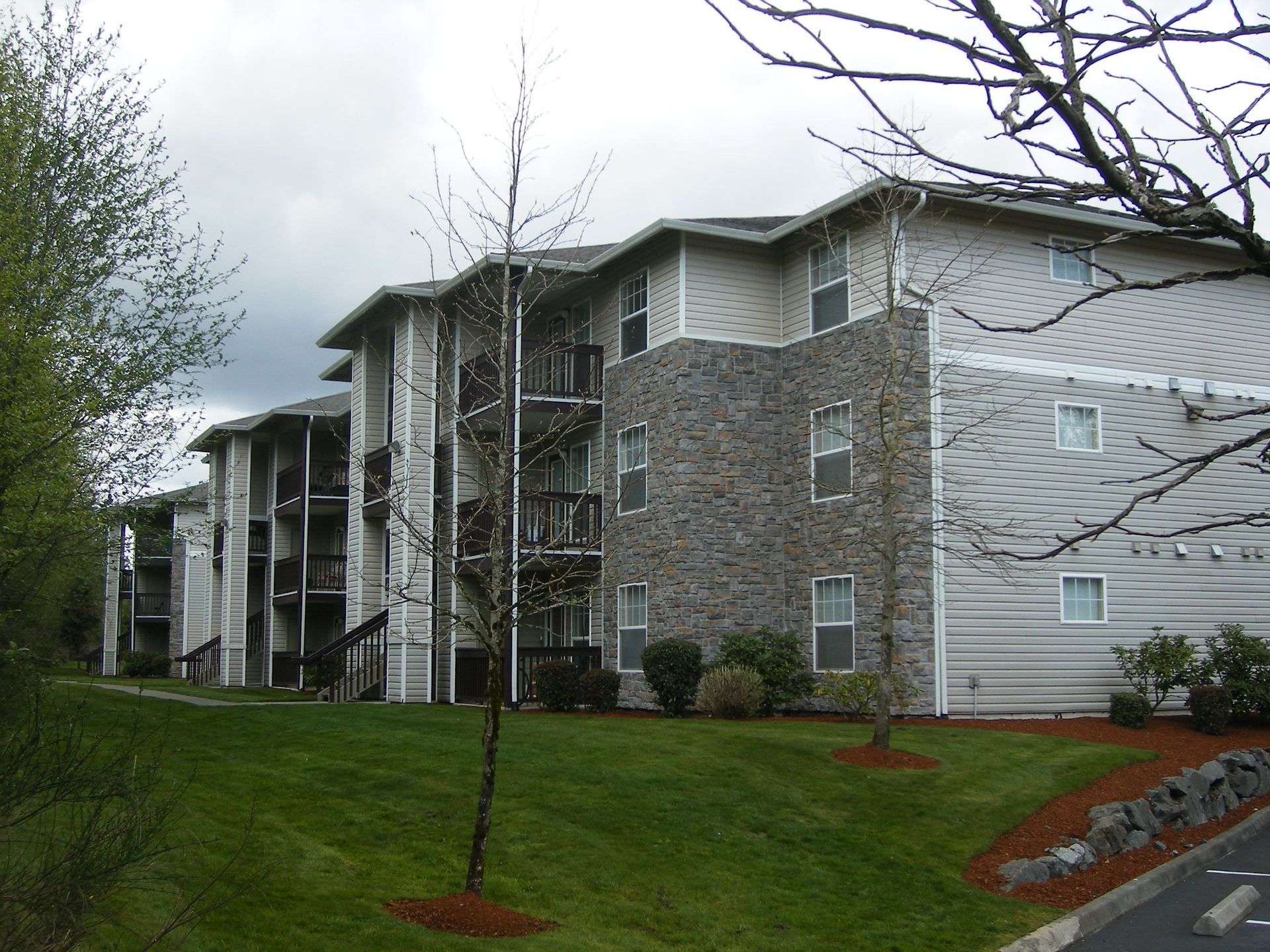 Apartment building with stone and white siding, seen beside a grassy lawn and small trees