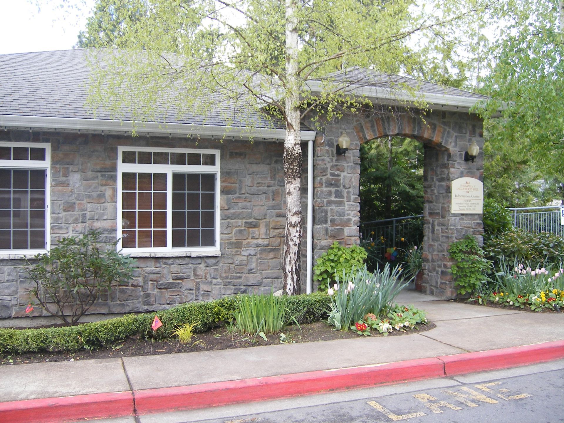 Stone building entrance with arched doorway, window, and landscaped flower bed by a red curb
