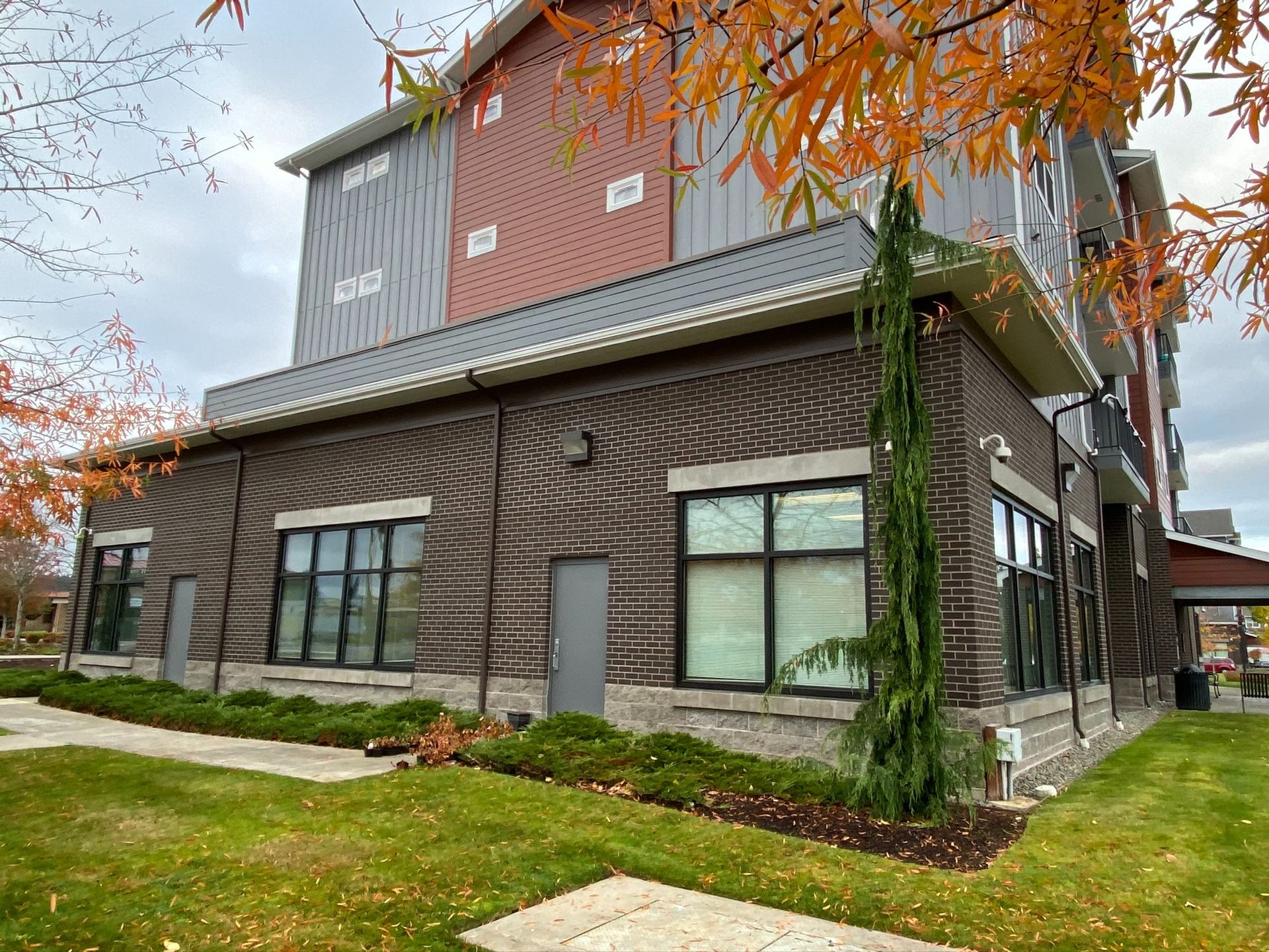 Modern two-story building with brown siding, large windows, and autumn trees beside a grassy lawn