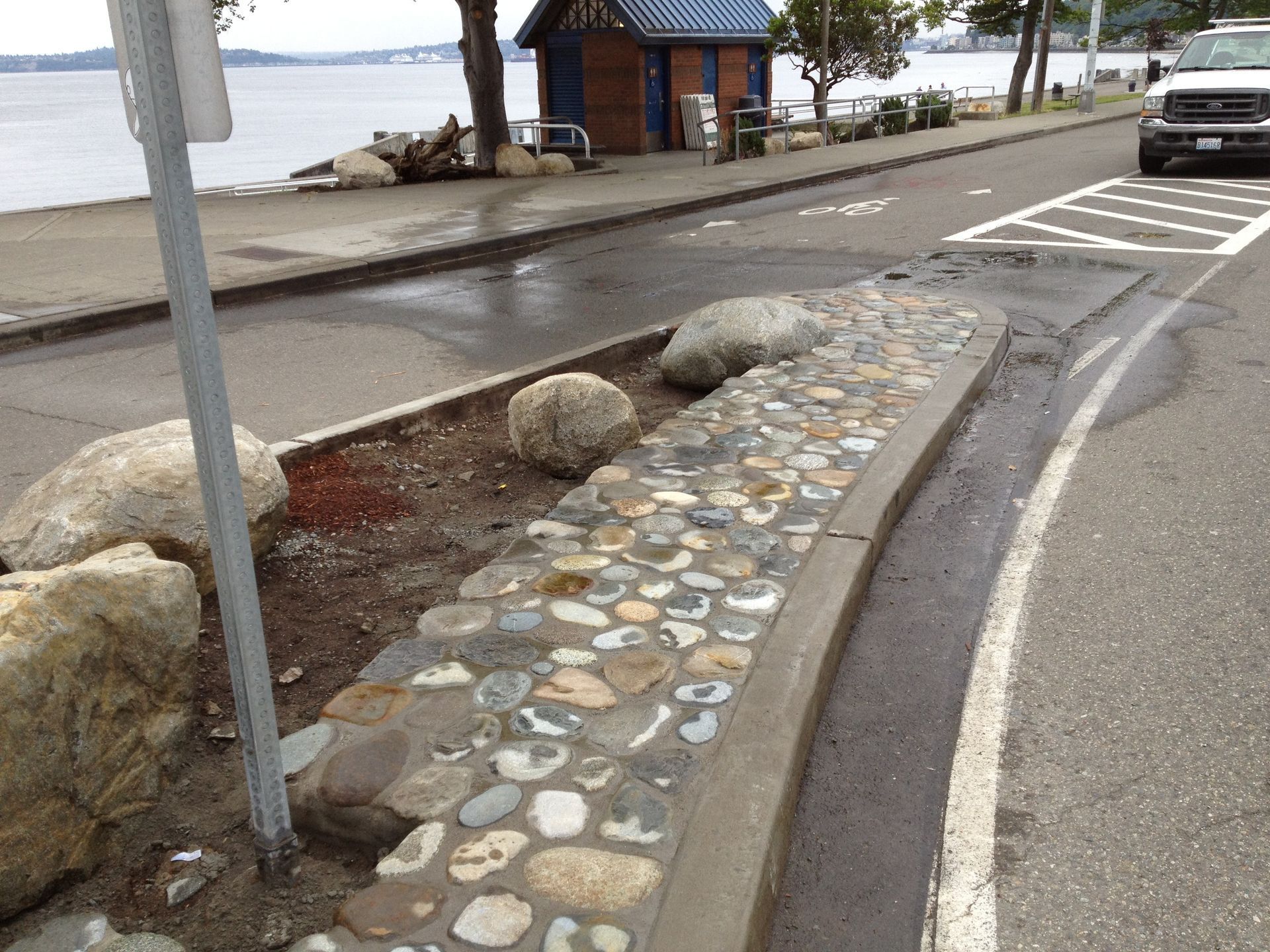 Curved stone-and-concrete walkway beside a waterfront road, with boulders and a parked car nearby.