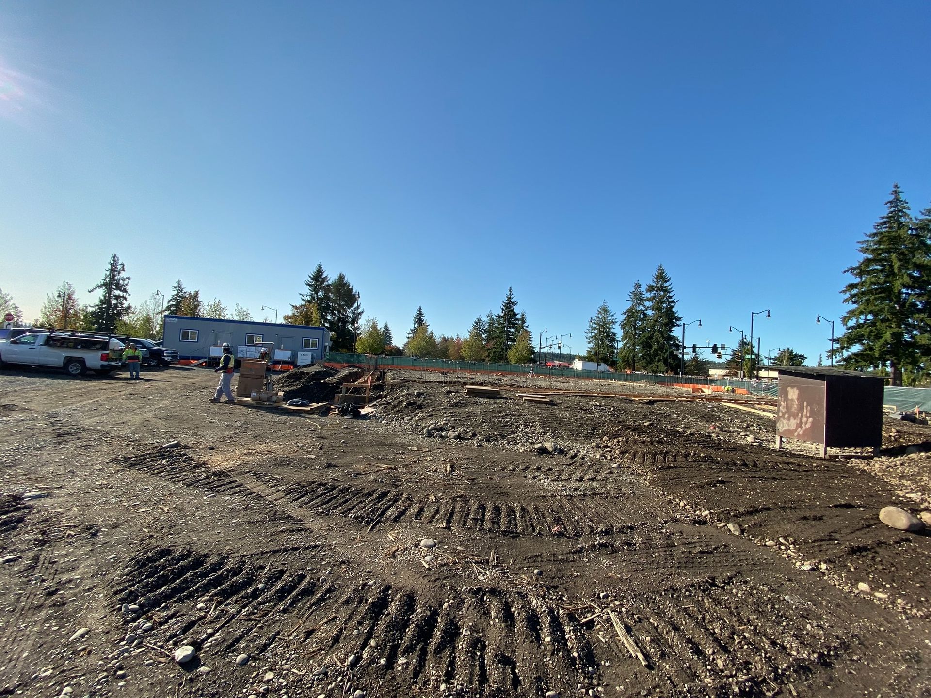 Gravel construction site with heavy equipment, dirt piles, parked vehicles, and tall trees under a clear blue sky
