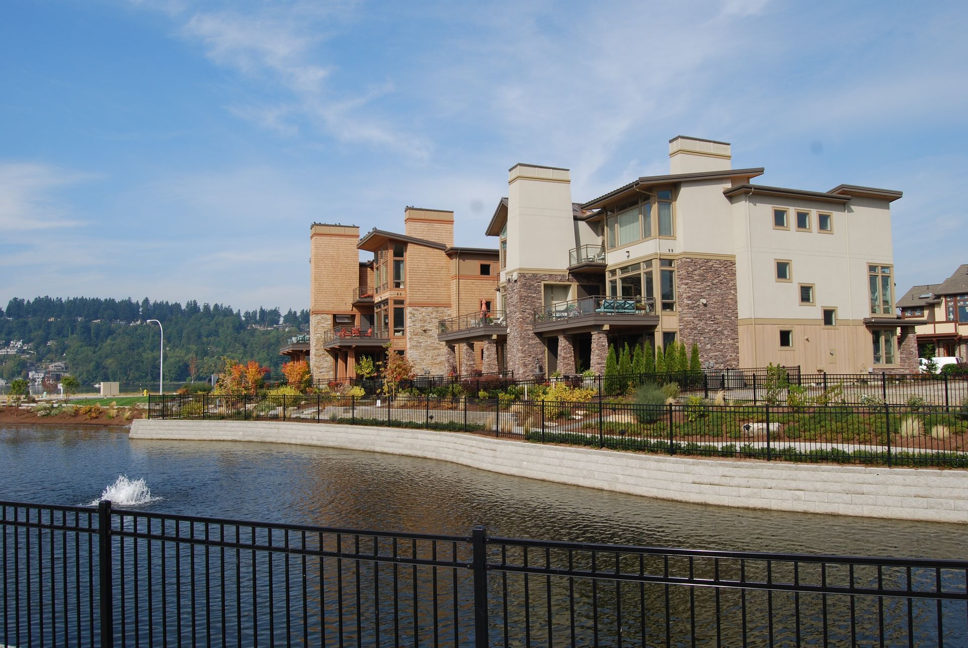 Waterfront homes and condos beside a calm bay, with a black fence in the foreground and trees in the distance