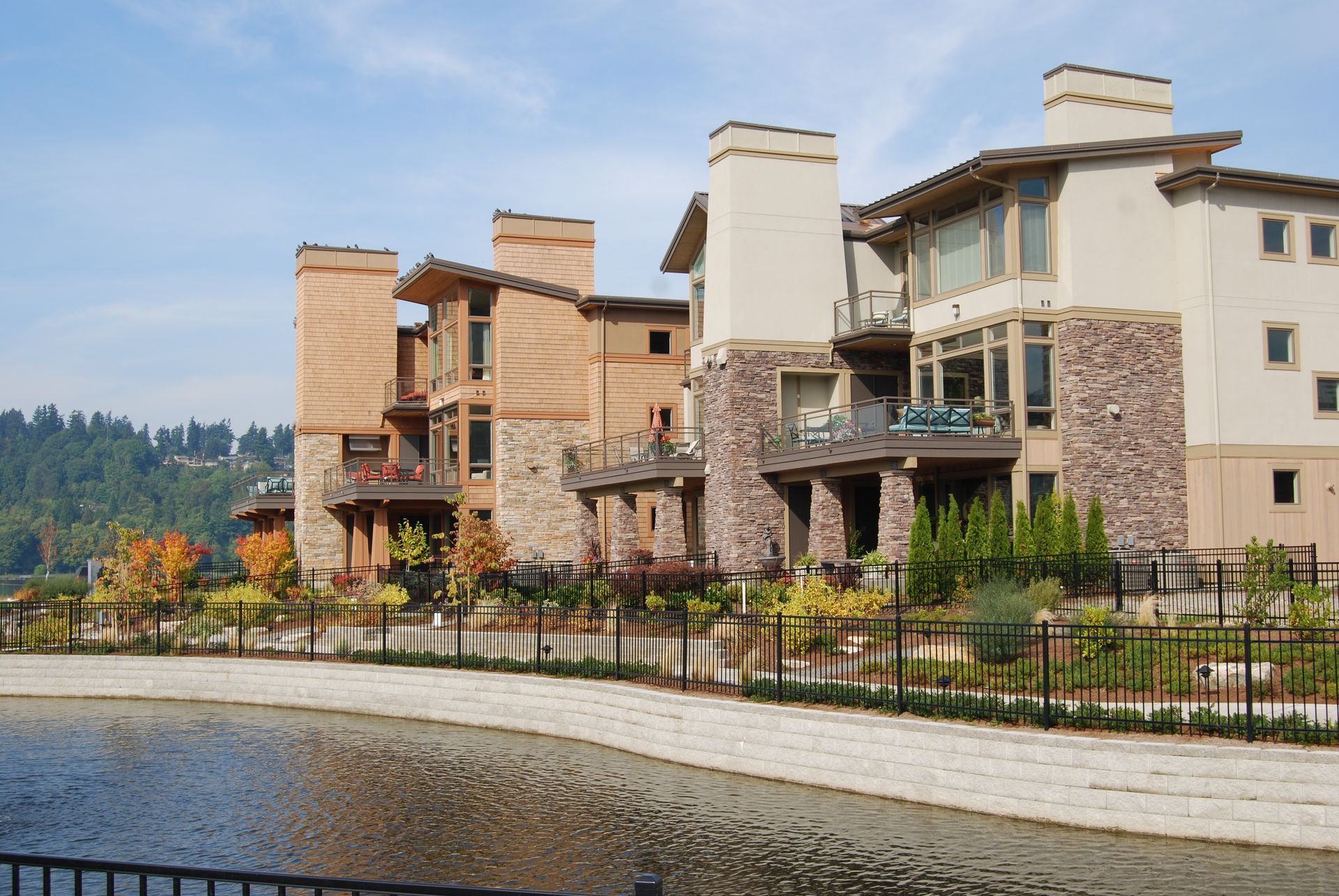 Waterfront row of modern stone and wood townhouses beside a canal, with trees and hills in the background.