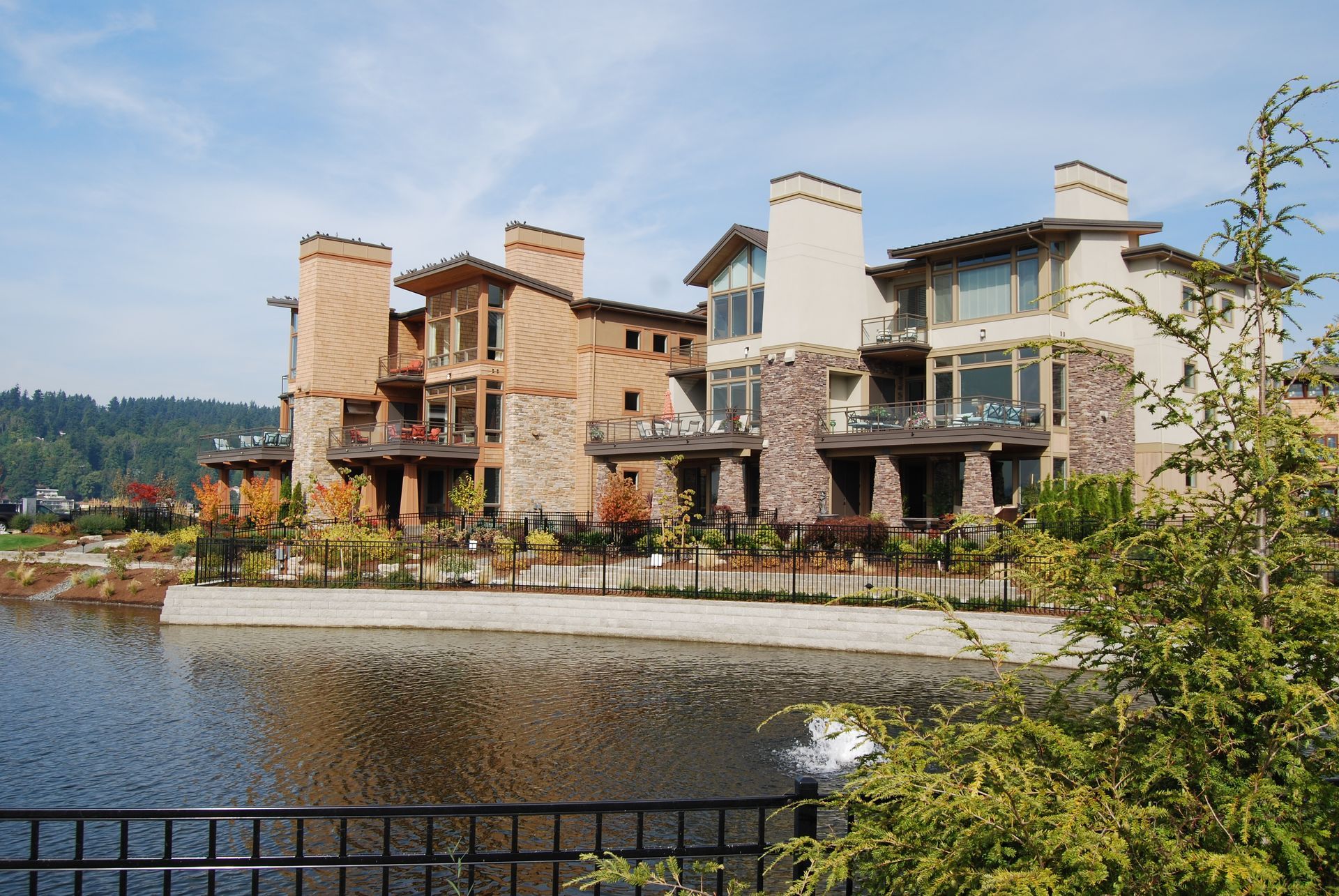 Large stone houses along a waterfront with a fenced path, autumn trees, and mountains in the background