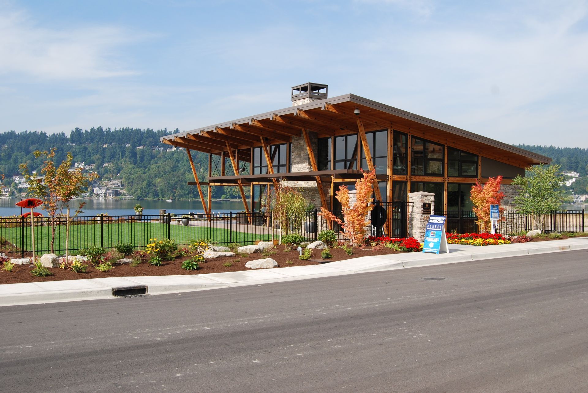 Modern building with a wooden roof, glass walls, and landscaped garden beside a road and lake.