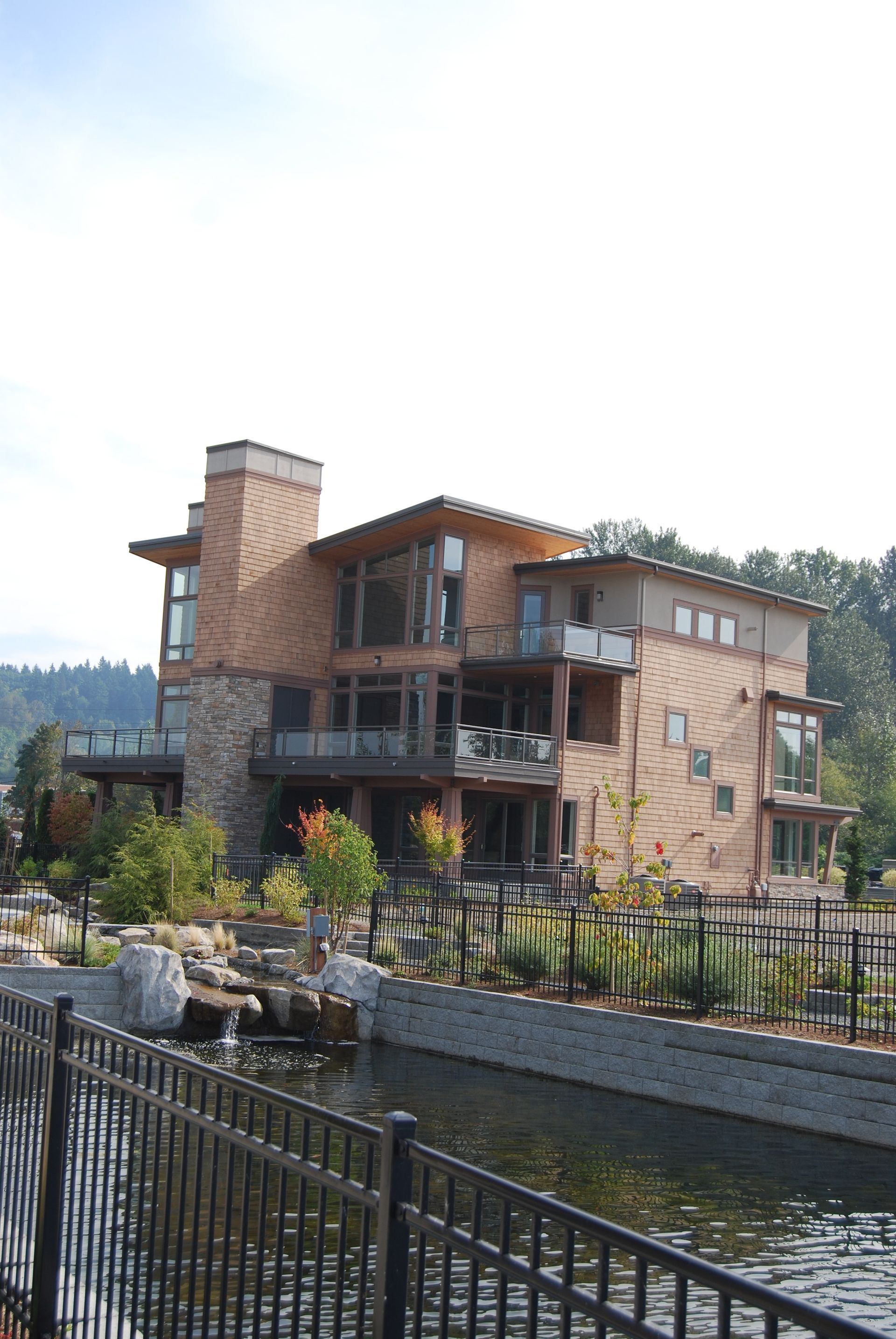 Modern brick-and-wood house beside a waterway with a black metal fence and landscaped garden