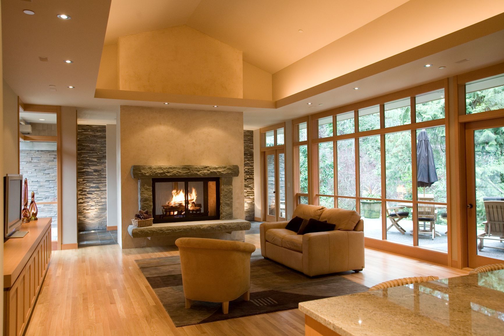 Modern living room with stone fireplace, tan seating, and large windows overlooking a wooded deck