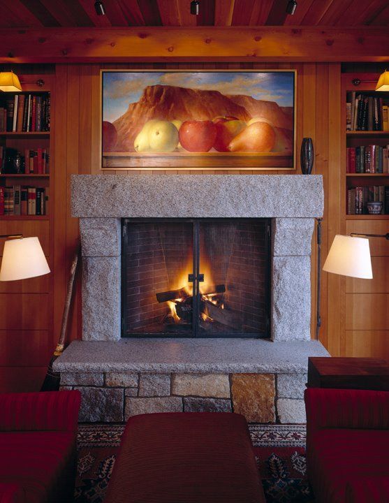 Cozy living room fireplace with a lit fire, stone mantel, wood-paneled walls, and a landscape painting above.