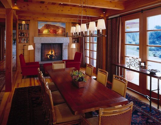 Warm dining room with a long wooden table, fireplace, red chairs, and large windows overlooking snow.