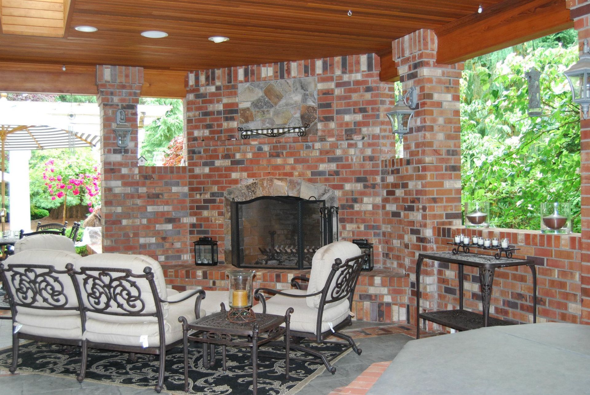 Covered brick patio with outdoor fireplace, cushioned seating, and wooden ceiling overlooking green trees