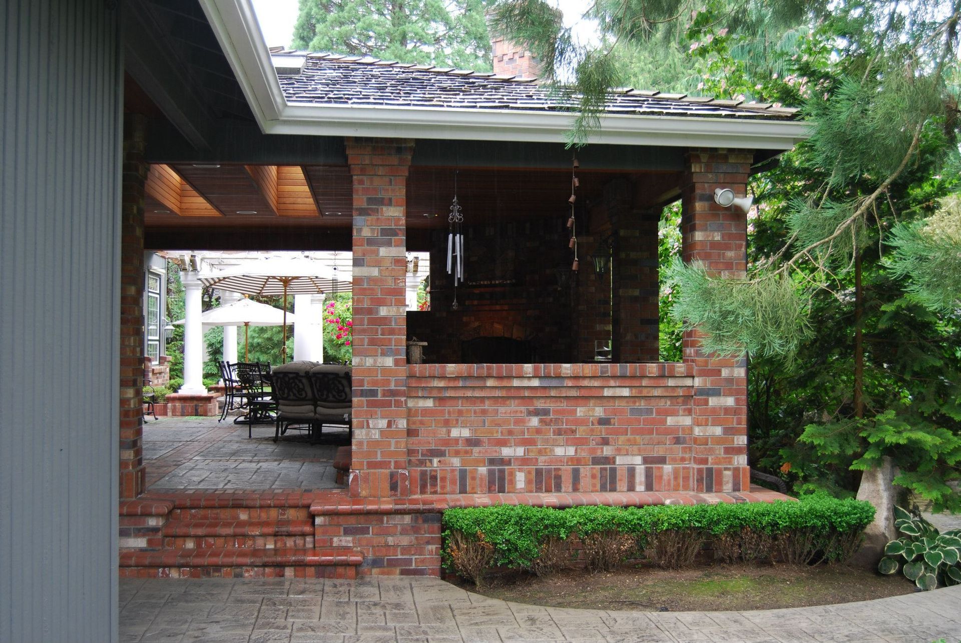 Brick porch with a screened seating area and green shrubs beside a house.