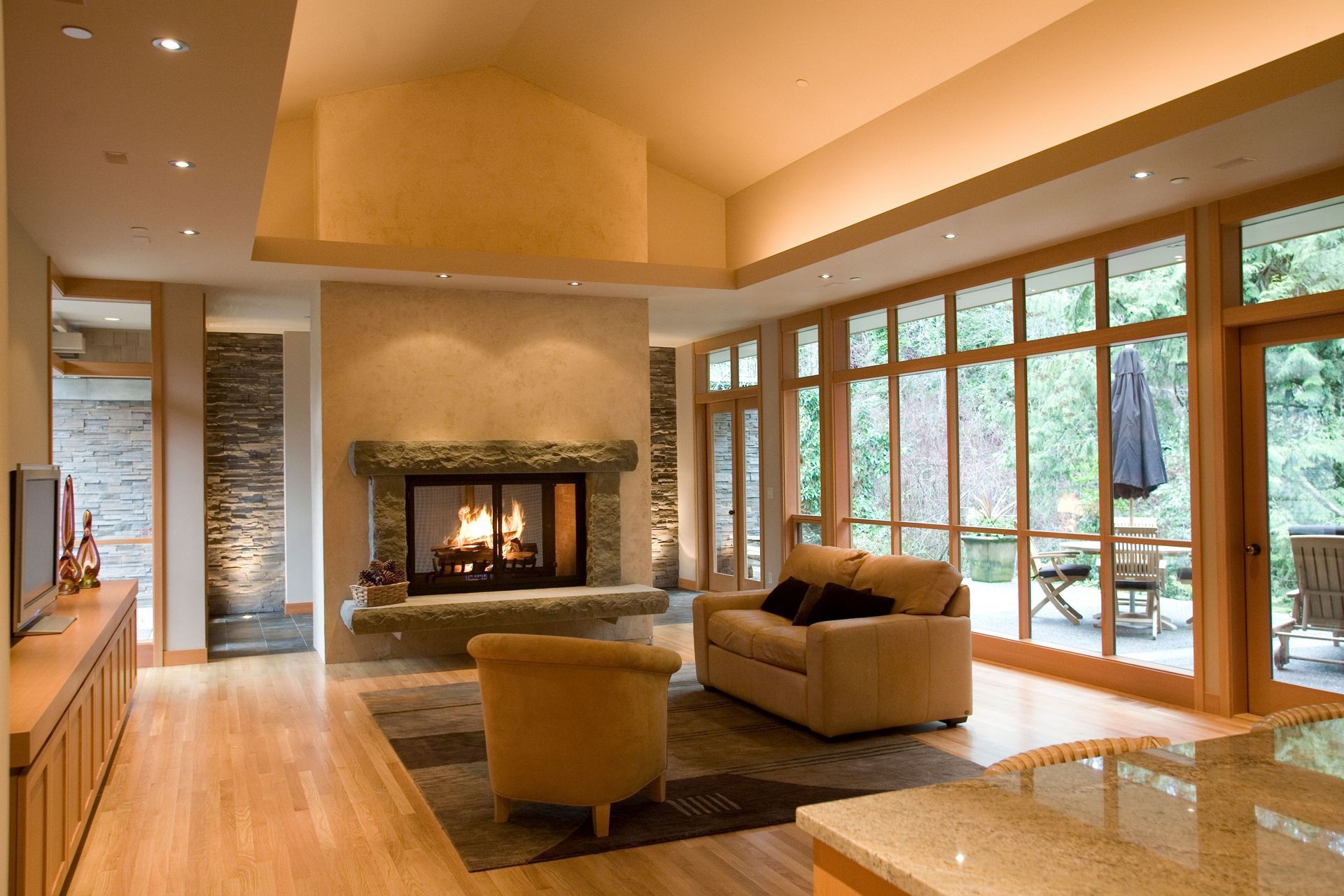 Modern living room with fireplace, tan seating, wood floors, and large windows overlooking greenery