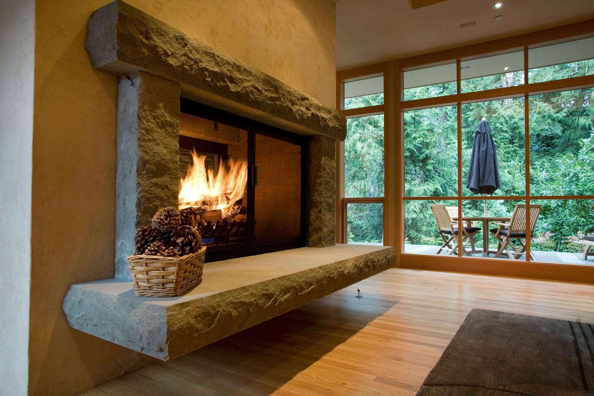 Cozy living room with a lit stone fireplace and large windows overlooking a wooded patio.