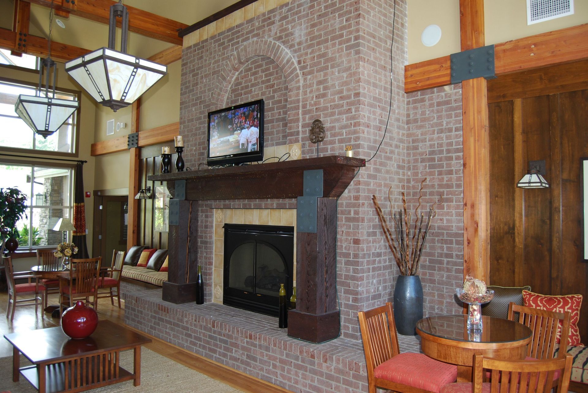 Living room with a brick fireplace, TV, and wood beams, featuring warm orange seating and large windows