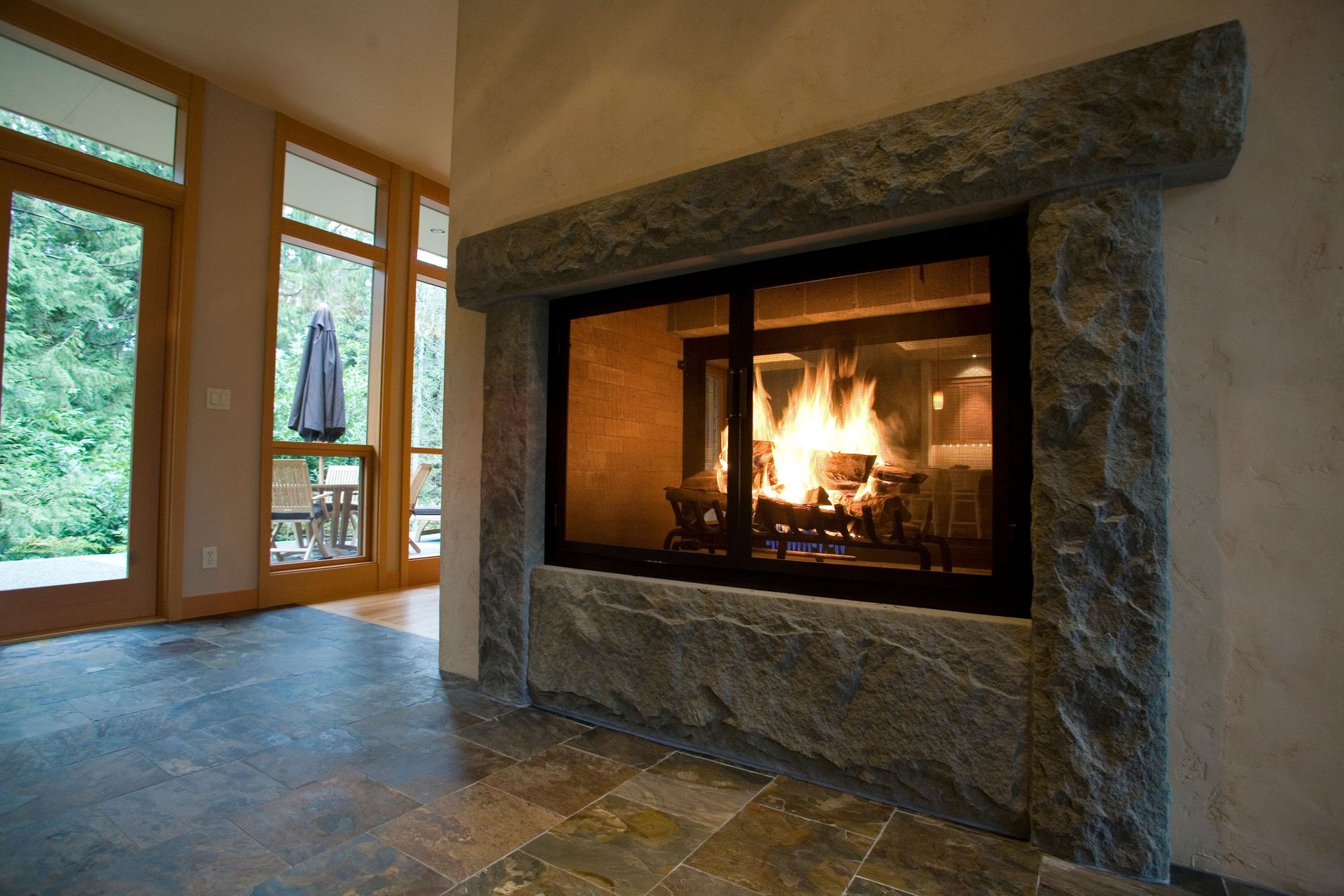 Stone fireplace with a burning fire in a bright living room with large windows and tile floor