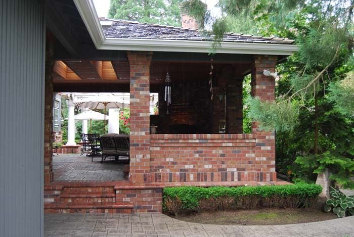 Brick patio and covered outdoor seating area beside a house, with brick columns and green shrubs.
