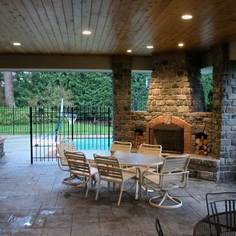 Covered patio with stone fireplace, white dining table, and view of a swimming pool beyond the fence