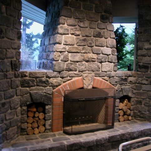 Stone fireplace with brick arch, stacked firewood, and windows overlooking trees