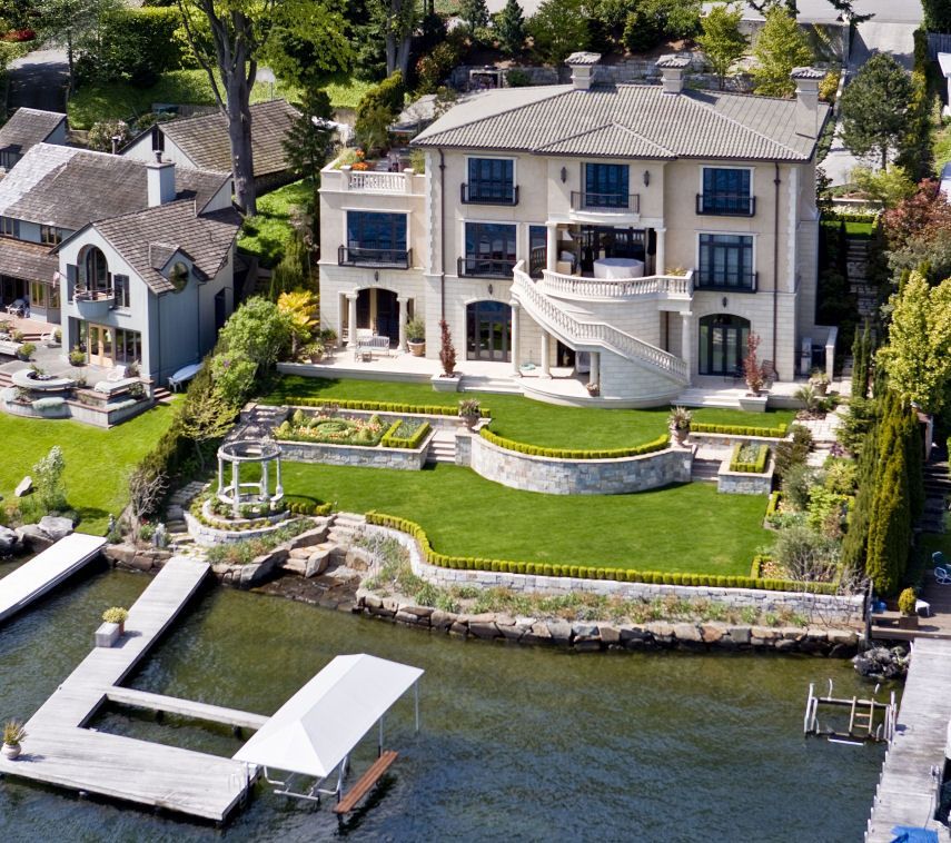 Luxury waterfront mansion with manicured lawn, stone terrace, and dock beside calm water
