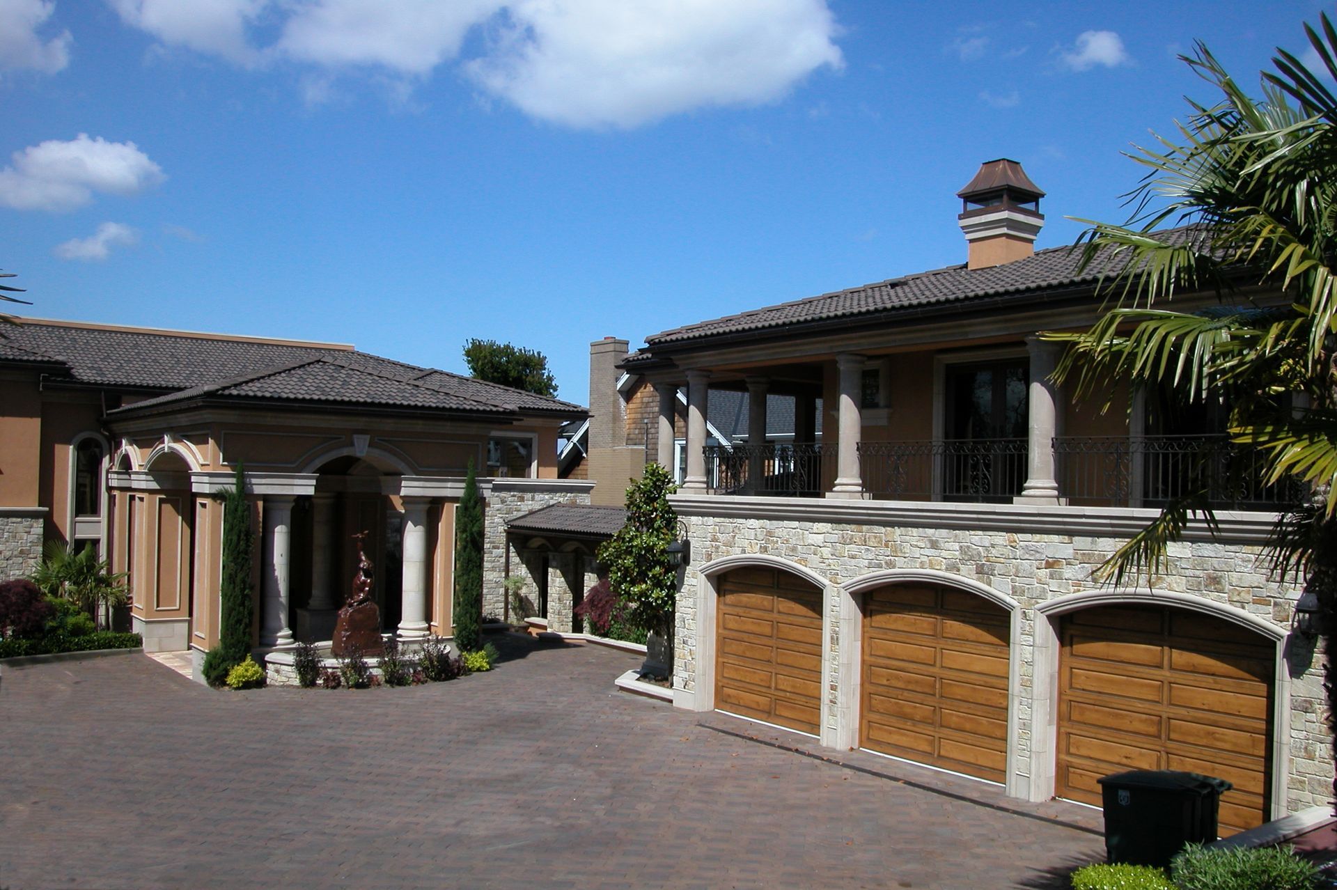 Large Mediterranean-style house with stone arches, wooden garage doors, and a paved driveway under a blue sky