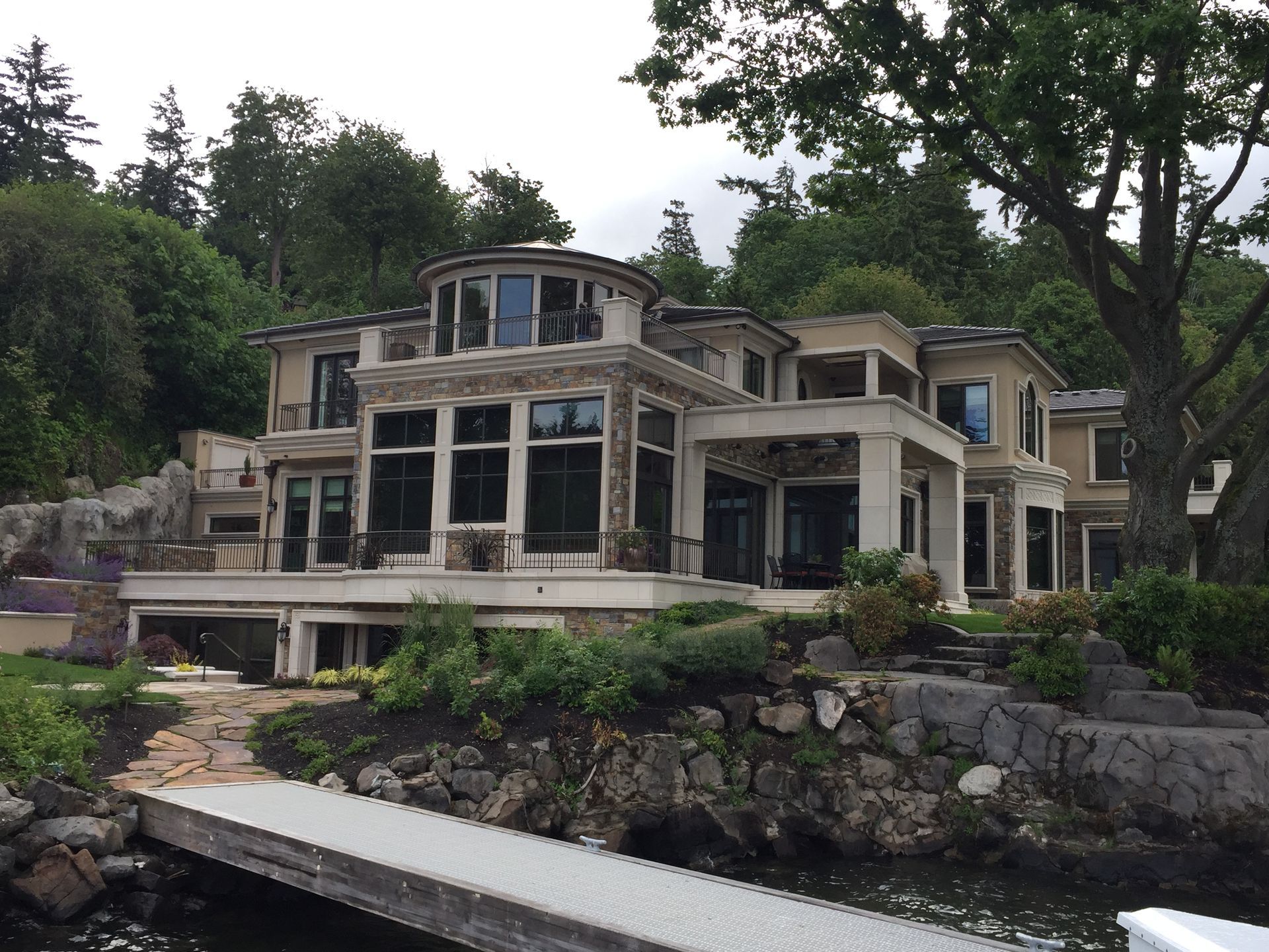 Large modern lakeside house with stone terraces and a dock, surrounded by trees and rocky shoreline