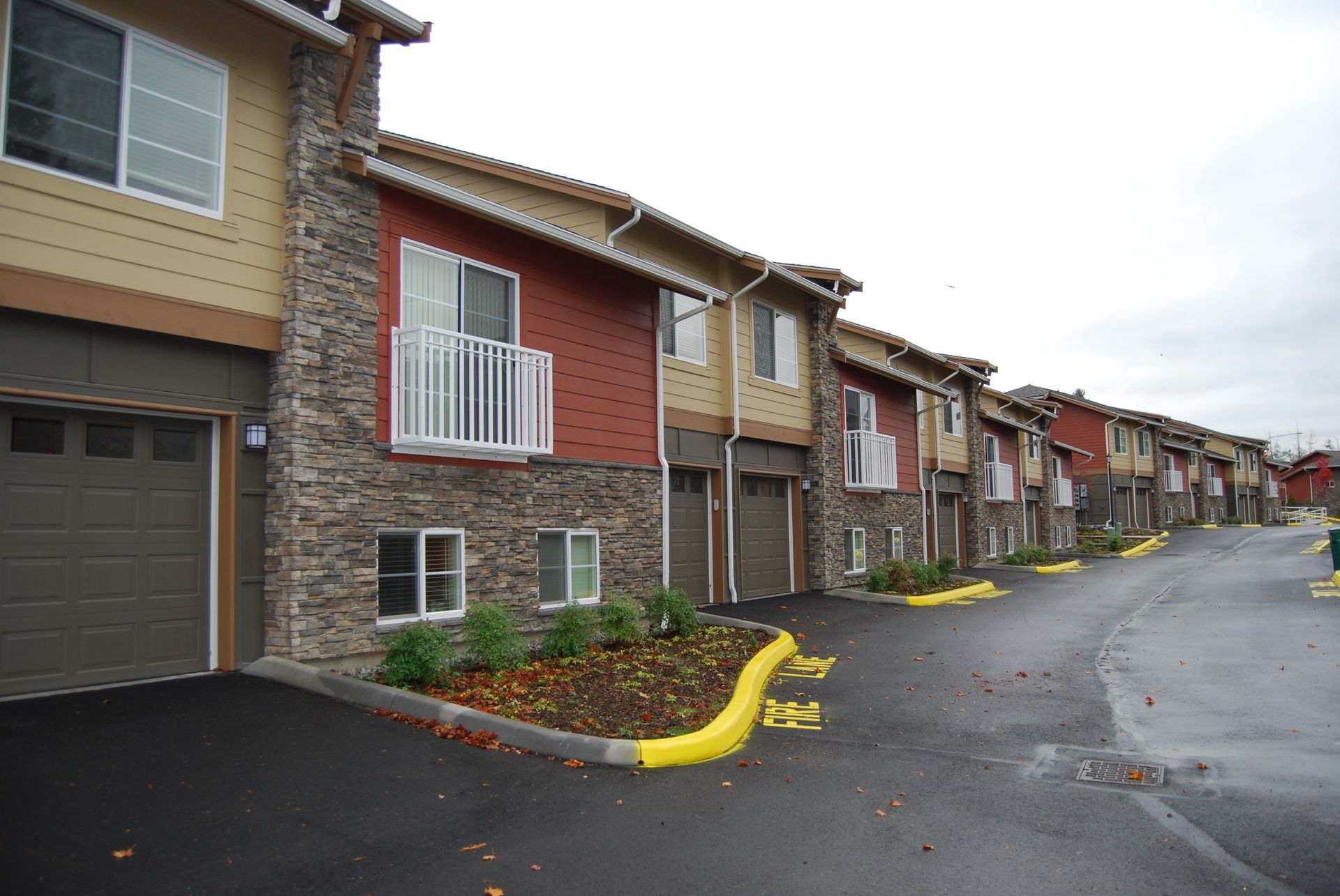 Row of beige and brick townhouses beside a wet driveway and yellow curbs on a rainy day