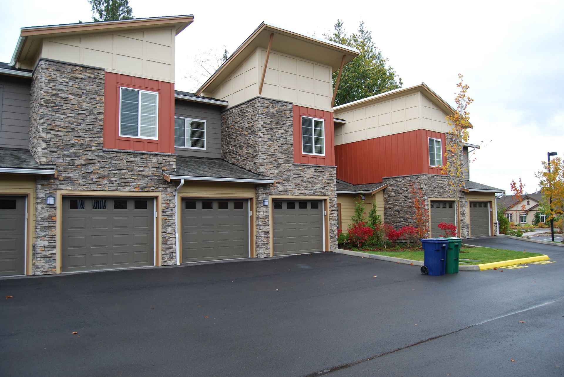 Modern townhouse row with stone and red siding, garage doors, driveway, and a blue trash bin