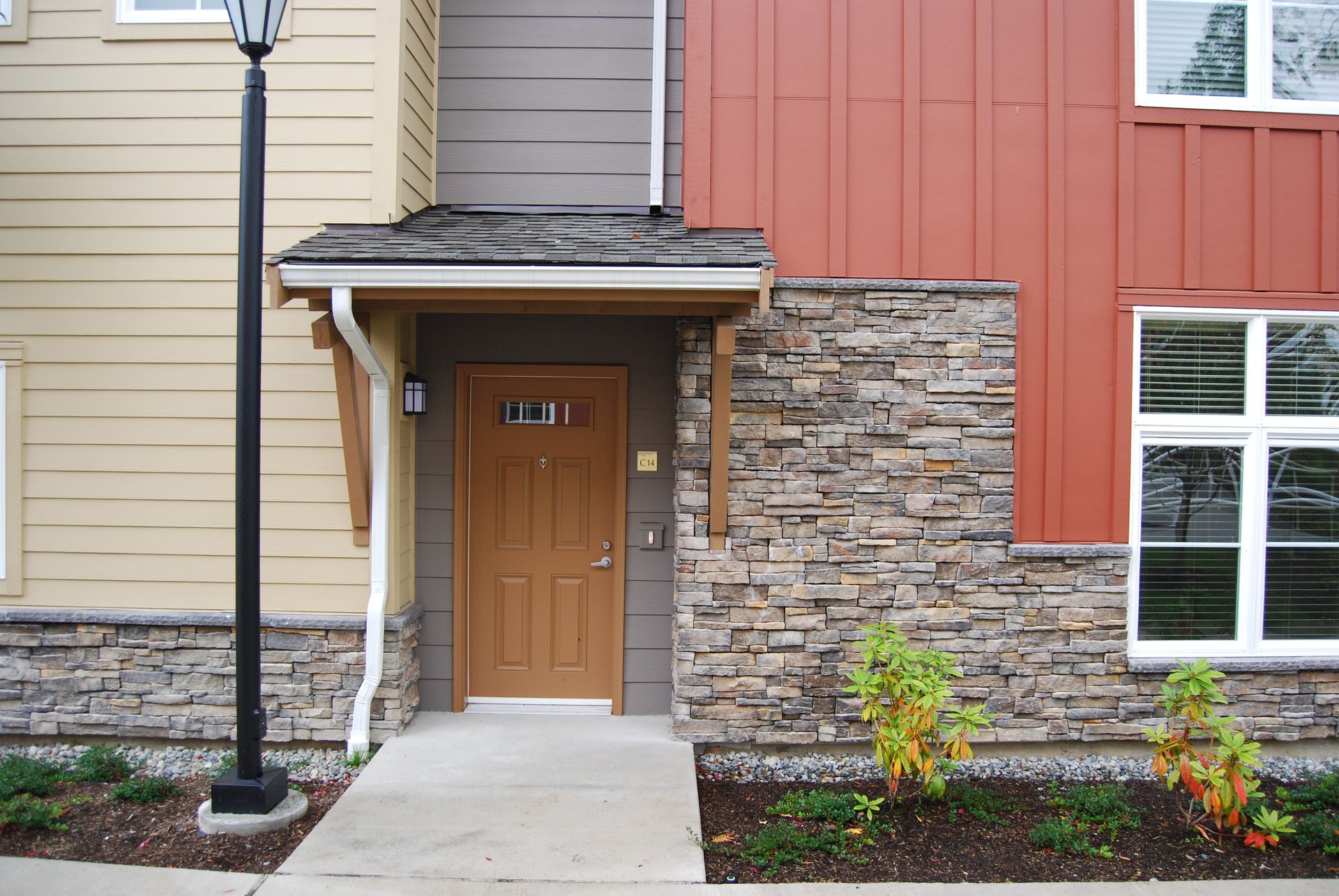 Apartment entrance with tan door, stone and beige siding, concrete walkway, and black lamp post