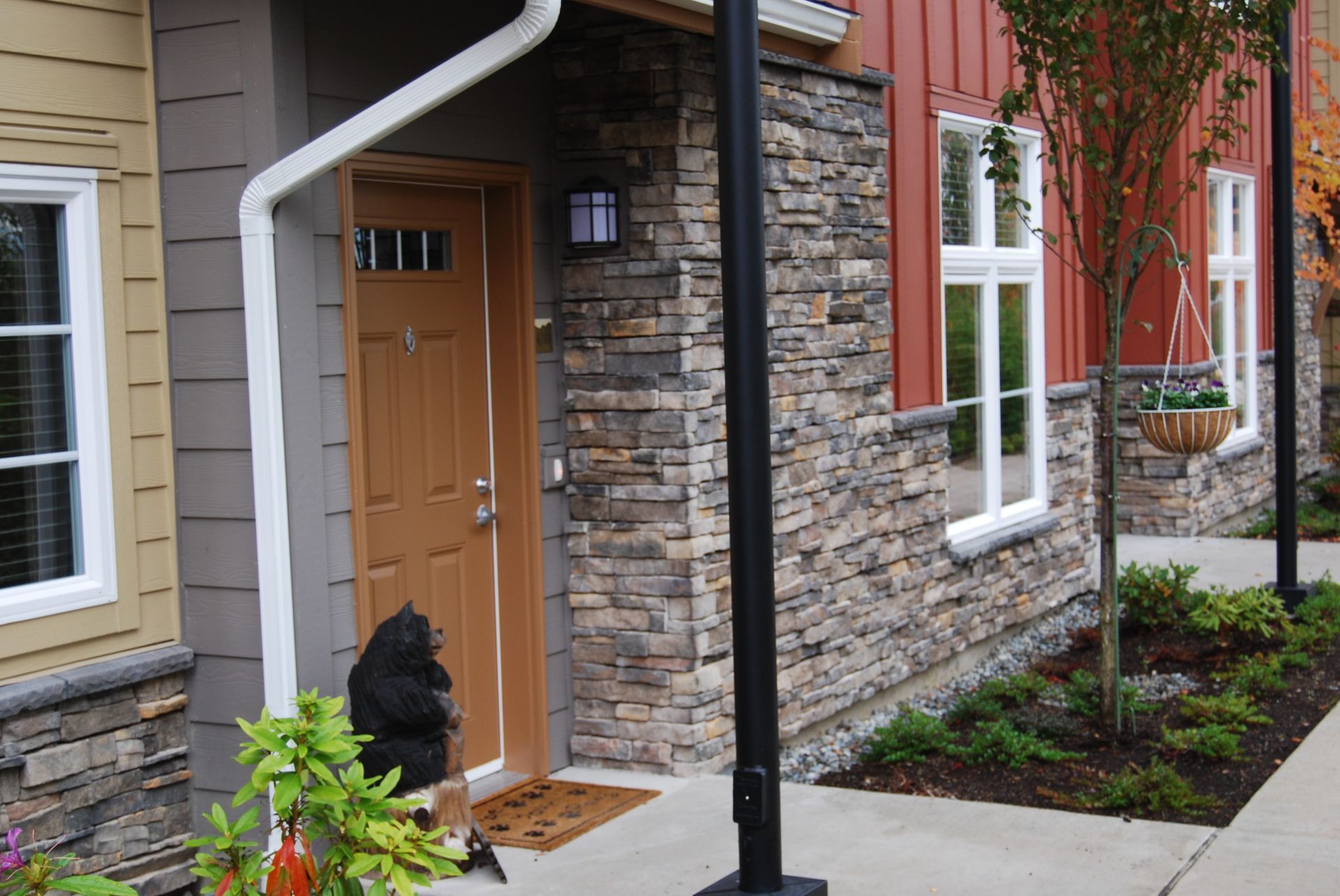 Front porch with tan door, stone and red siding, black lamp post, and small garden beds.