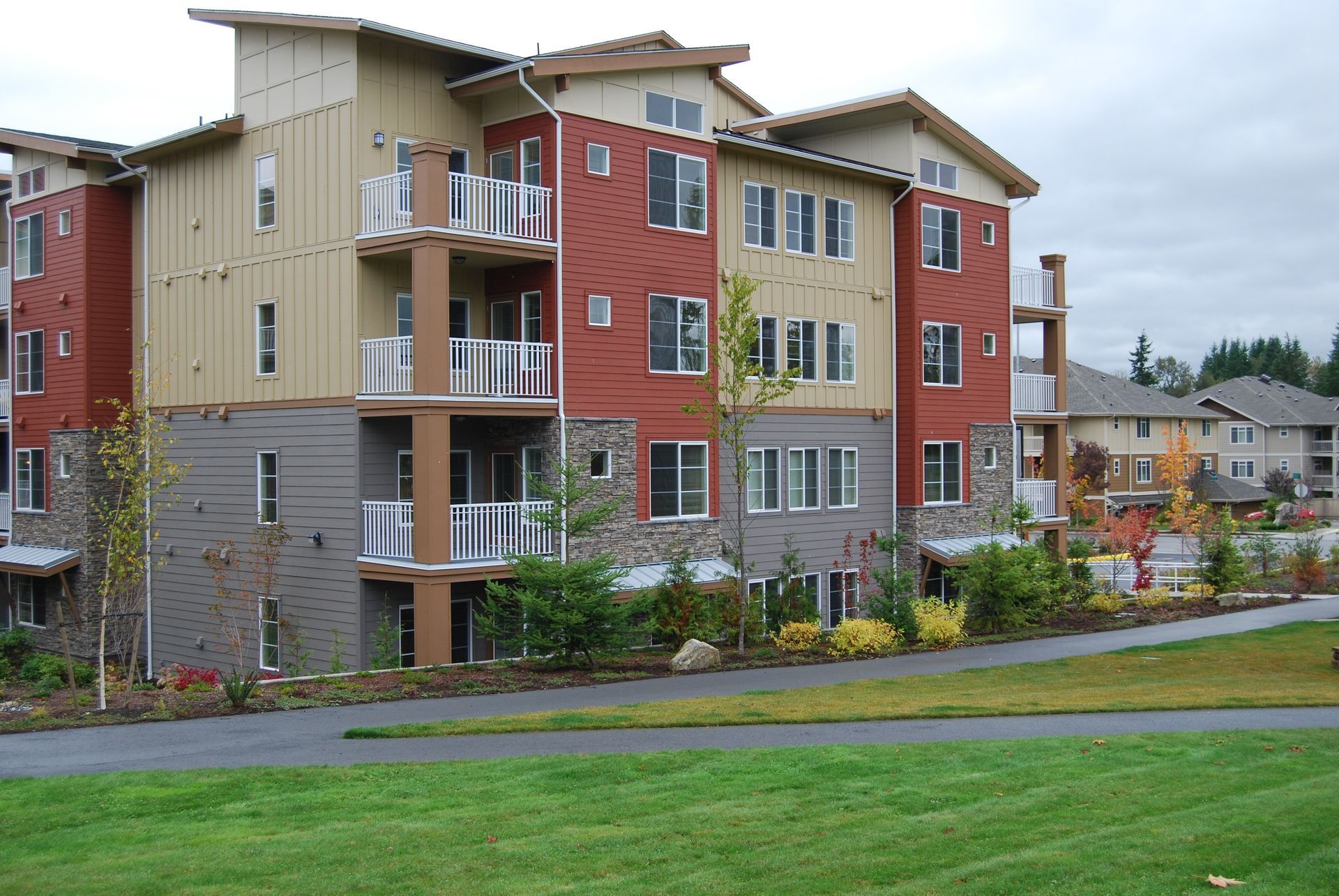 Modern apartment building with red, tan, and gray exterior beside a green lawn and cloudy sky