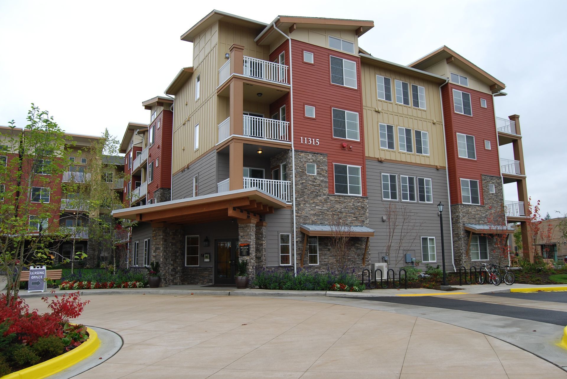 Modern multi-story apartment building with red and beige exterior, stone base, and covered entrance on a curved driveway