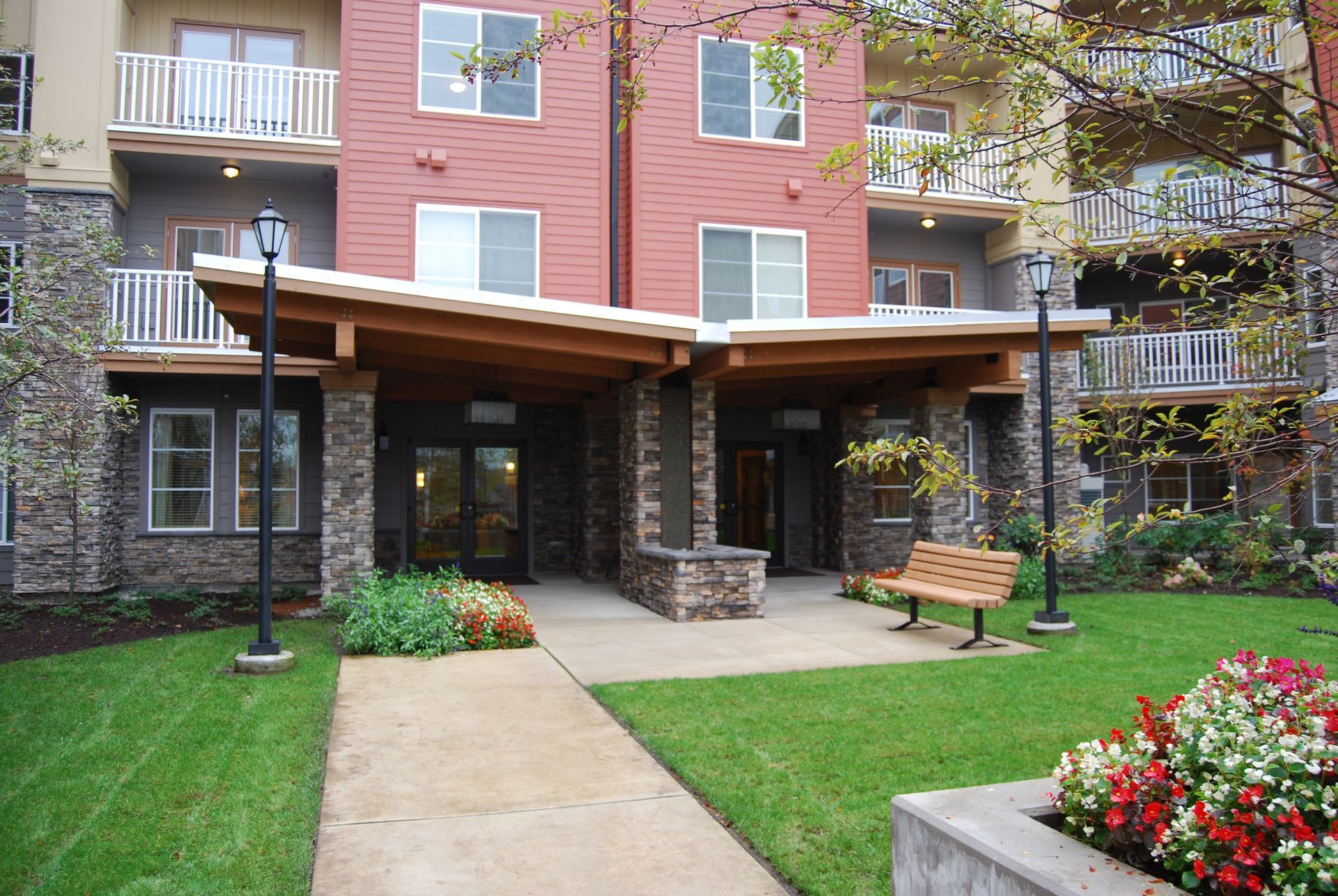Courtyard entrance with stone pillars, wooden canopy, bench, walkway, and landscaped flowers beside apartment buildings