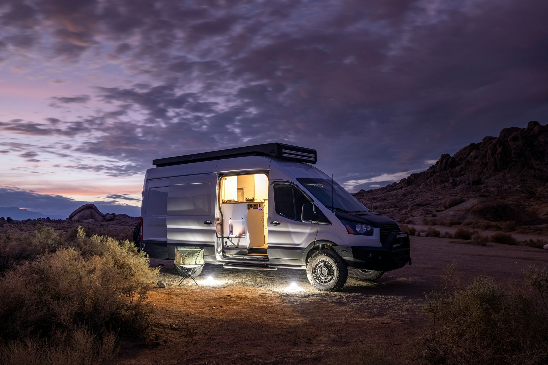 Camper van parked in a desert at dusk, with warm lights glowing inside and outside.