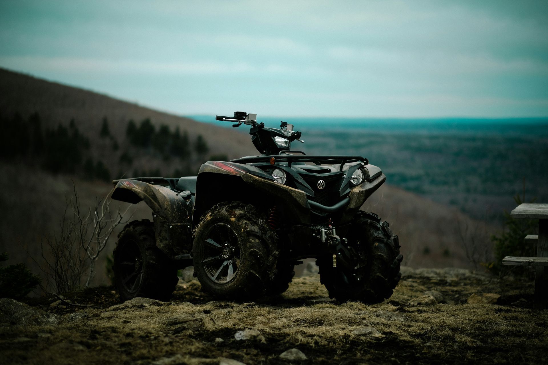 Black ATV parked on a rocky hilltop overlooking a cloudy forested landscape.