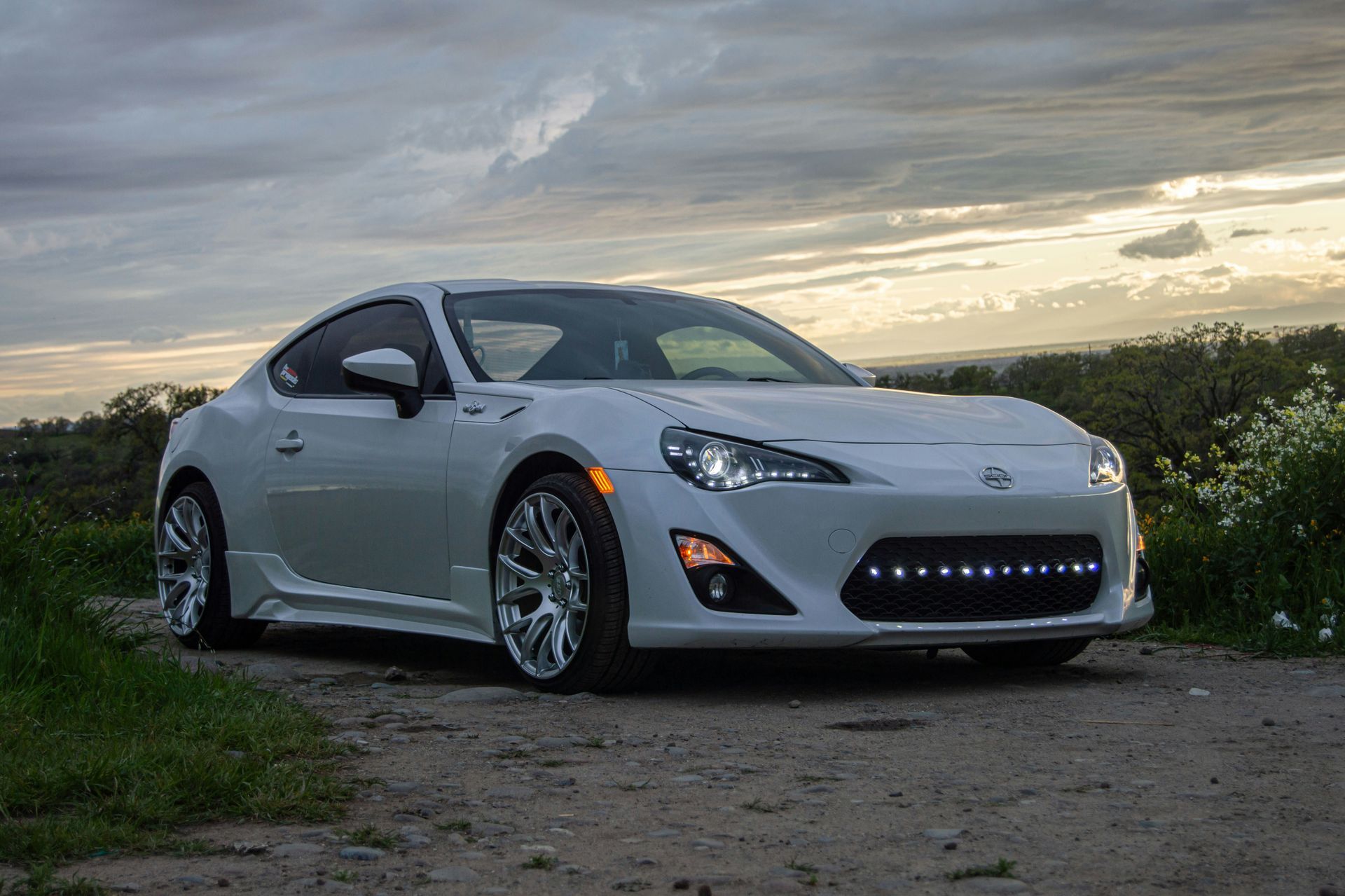 White sports car parked on gravel at sunset with a cloudy sky behind it