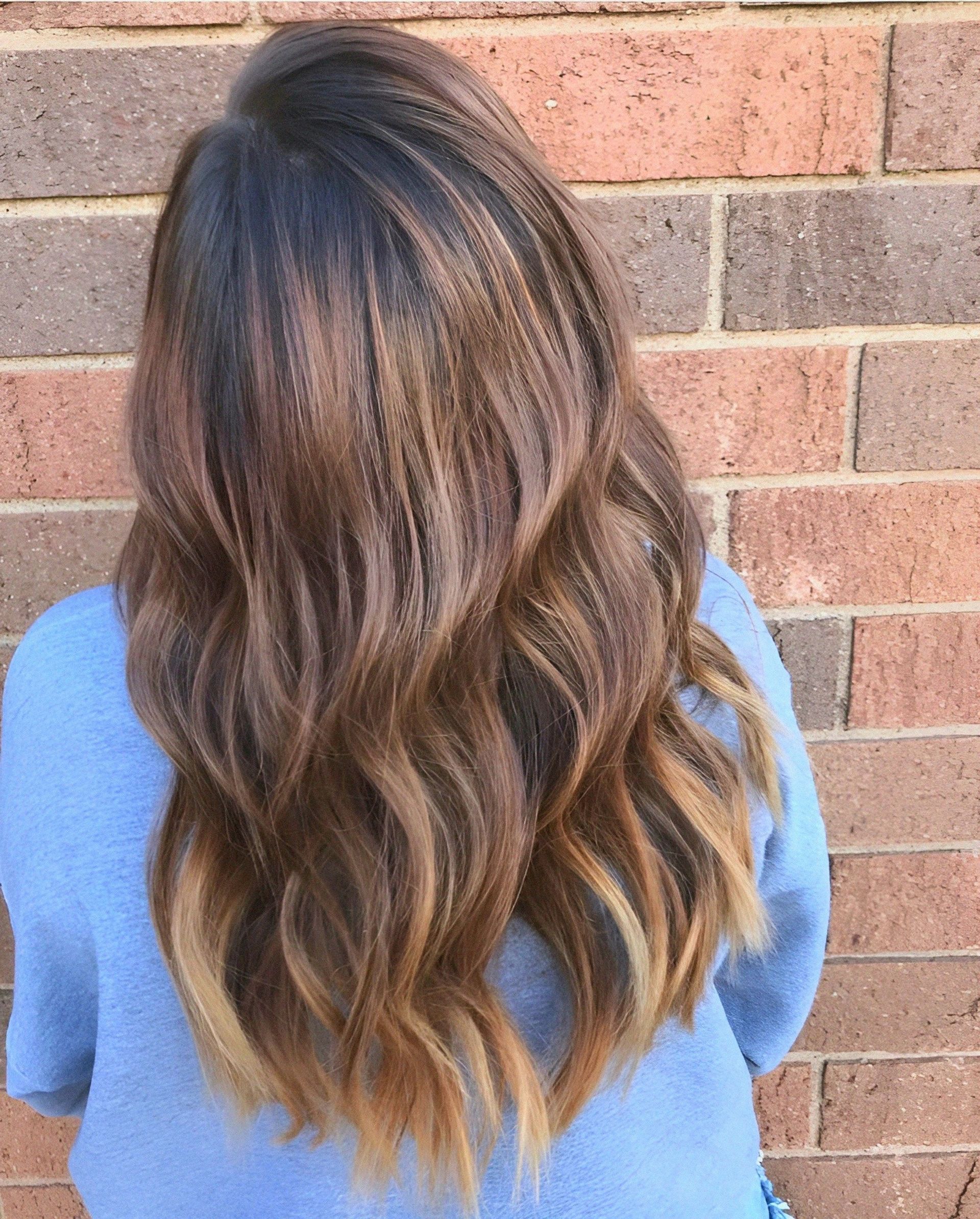 Woman with long wavy brown hair, transitioning to blonde ends, in front of a brick wall.