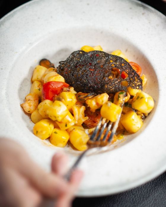 Primo piano di una ciotola bianca con gnocchi, cozze, pomodori e un piatto scuro e incrostato. Una mano regge una forchetta.
